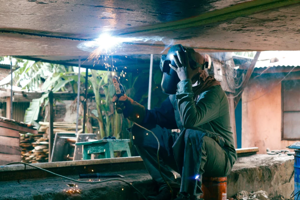 Worker performing overhead welding with protective gear and helmet in an outdoor workshop, sparks flying during metal fabrication.